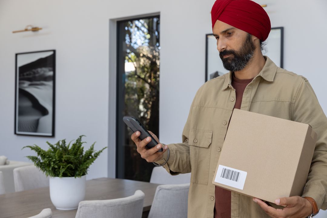 Man with Turban Holding Shipping Box and Using Smartphone at Home