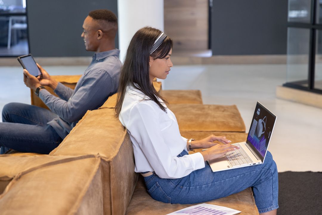 Young diverse coworkers using laptop and tablet on lounge couch for remote collaboration