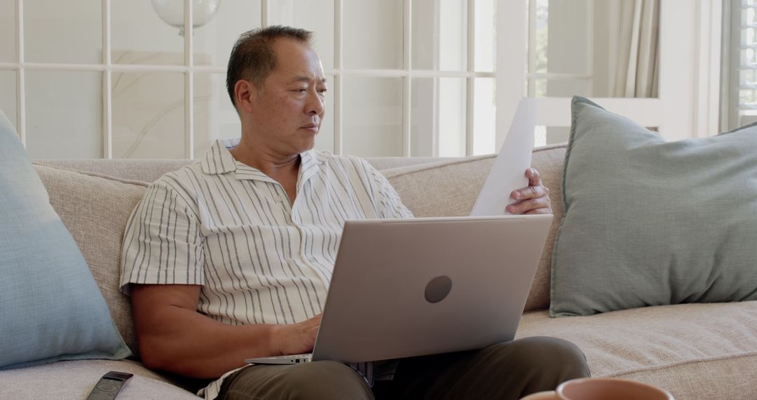 Asian Man Analyzing Documents and Working on Laptop at Home