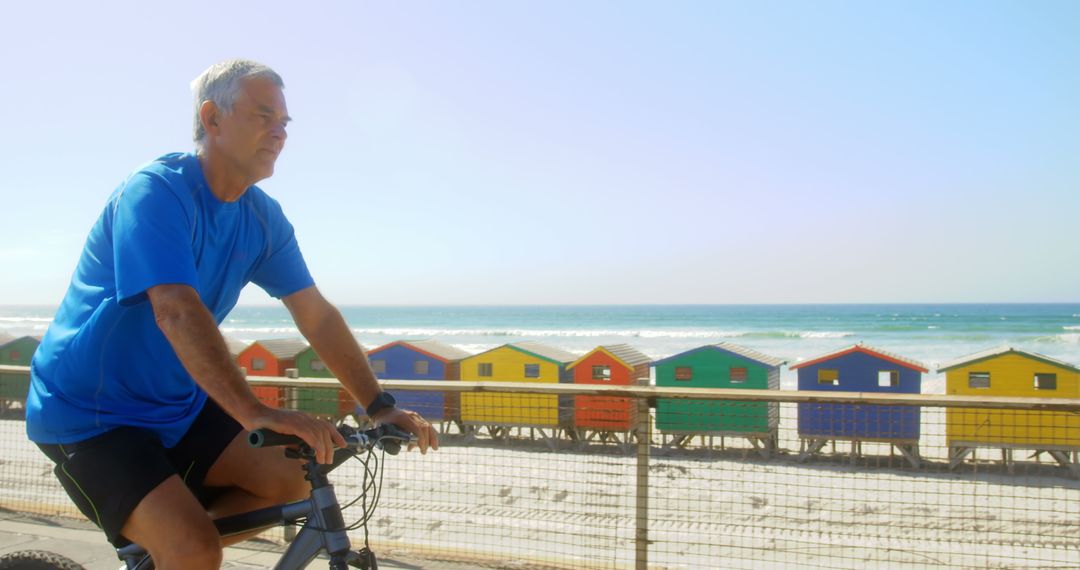 Senior Man Biking by Beachfront with Colorful Huts in Background