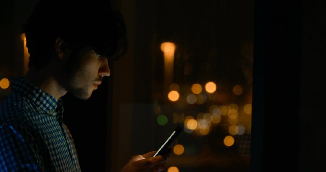 Young man staring at smartphone by window at night with city bokeh and reflective glass