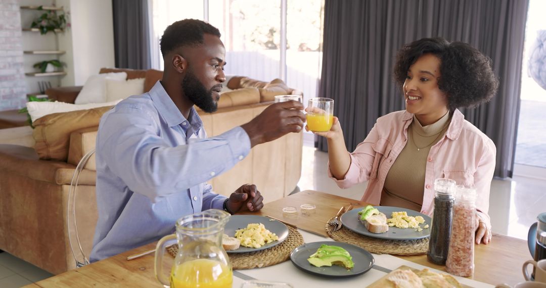 Black couple toasting with orange juice at cozy sunlit breakfast table, avocado toast