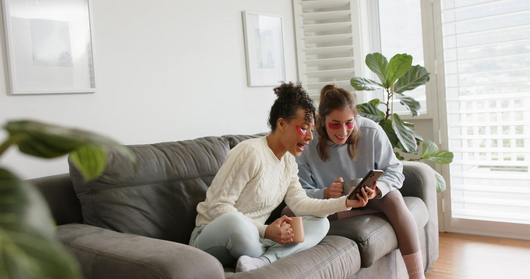Two Friends Sharing Self-Care Morning with Under-Eye Patches on Cozy Sofa, Browsing Phone
