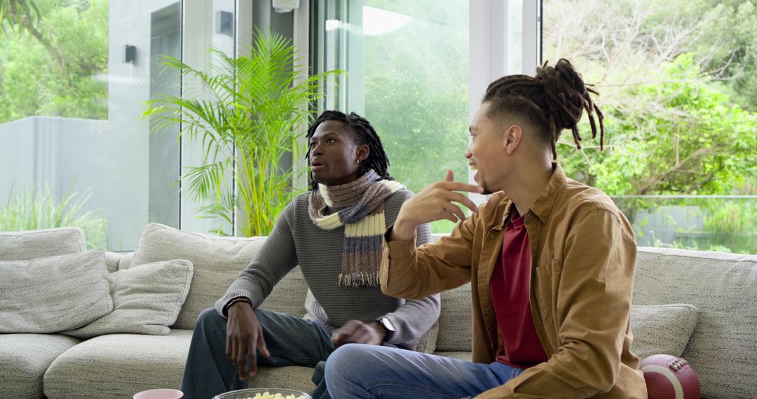 Diverse male friends chatting on sofa with popcorn and football in sunlit home