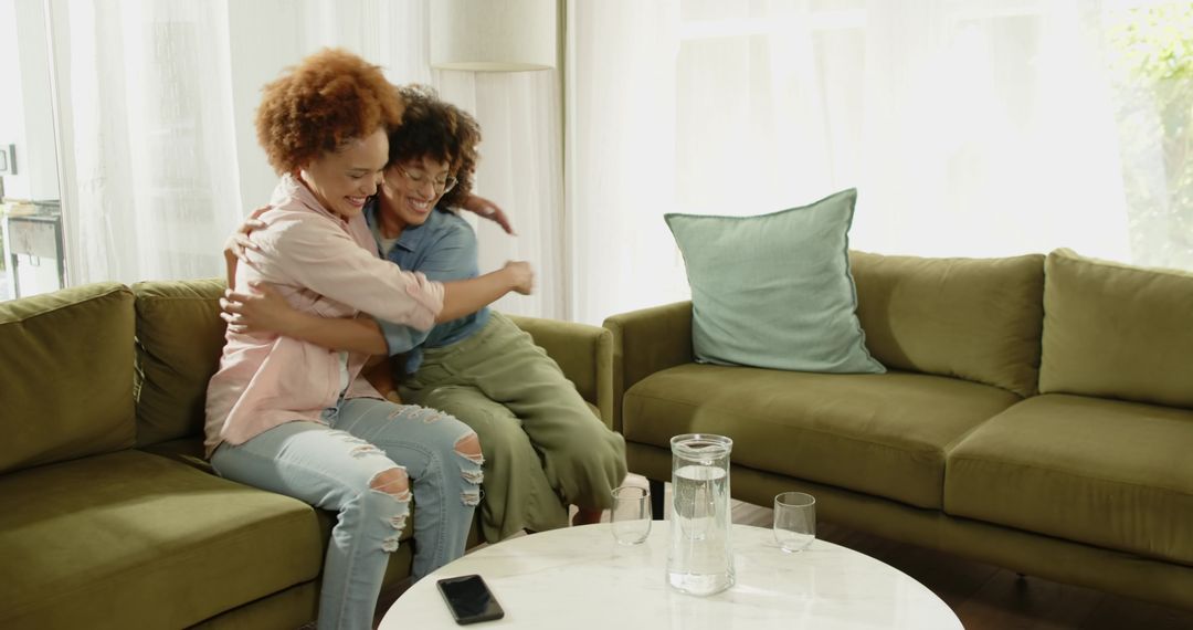 Two friends hugging and smiling on olive-green sectional sofa in sunlit living room