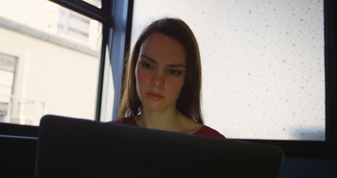 Focused Woman Working on Laptop Near Window in Office - Free Images, Stock Photos and Pictures on Pikwizard.com