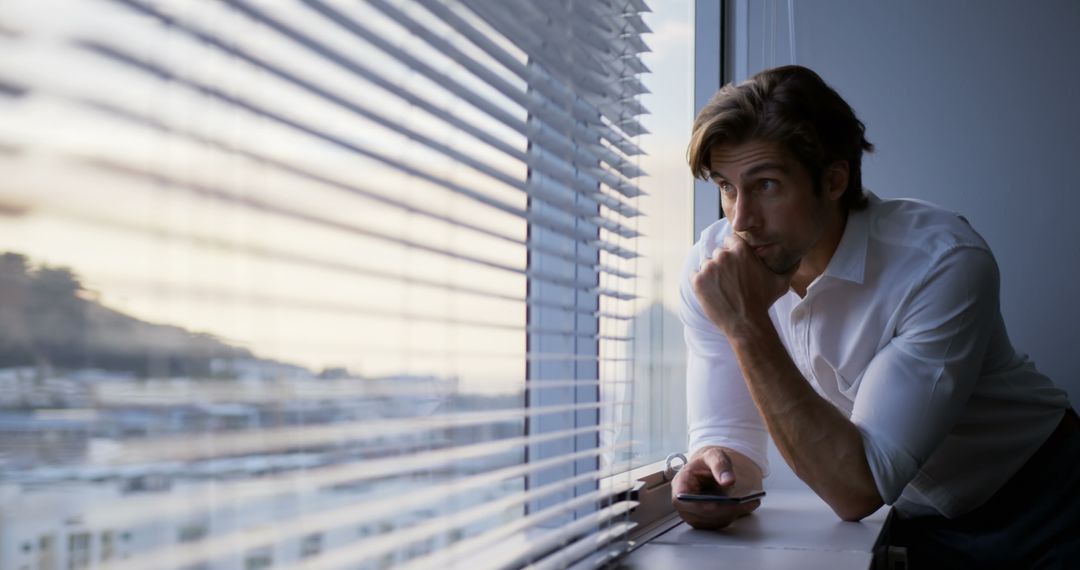 Pensive Businessman Looking Through Office Window with Smartphone