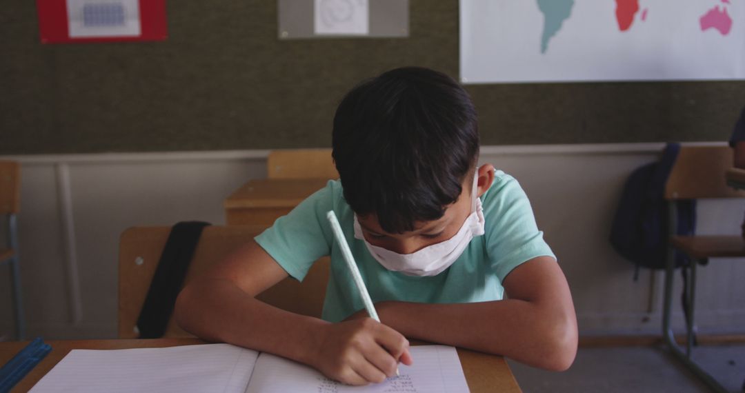 Diverse boy sitting at desk, wearing facemask and writing in notebook in elementary school class - Free Images, Stock Photos and Pictures on Pikwizard.com