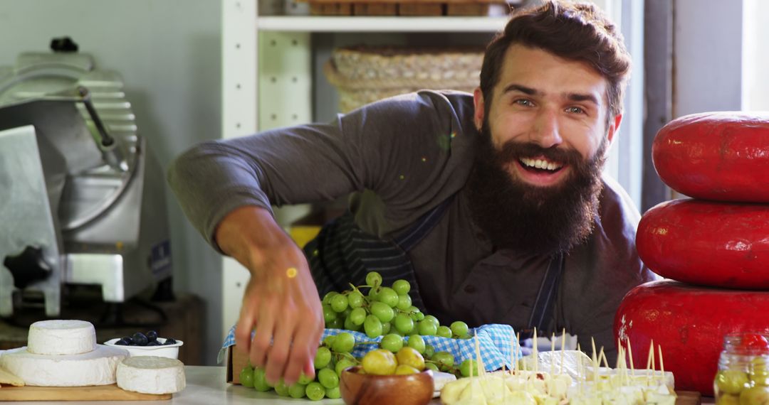 Cheerful Bearded Man Arranging Fresh Cheeses and Grapes in Delicatessen - Free Images, Stock Photos and Pictures on Pikwizard.com