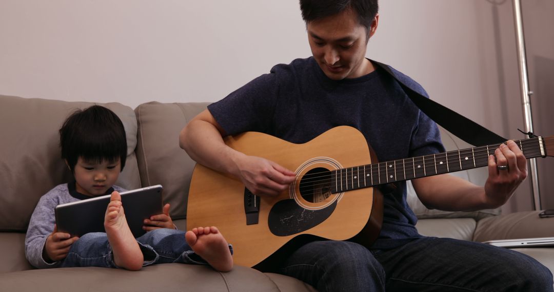 Father Playing Guitar While Child Uses Tablet on Couch - Free Images, Stock Photos and Pictures on Pikwizard.com