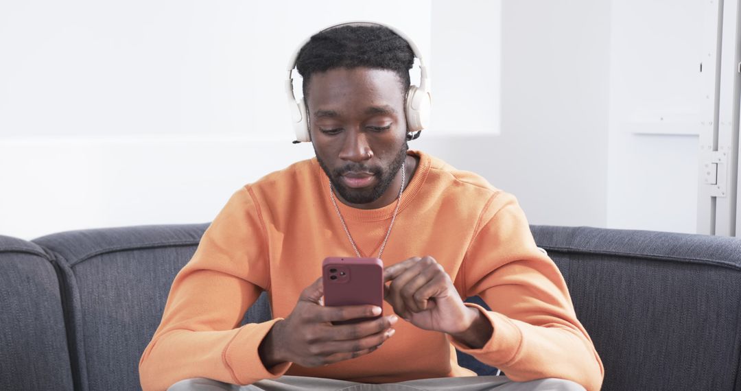 African-American man using smartphone while wearing headphones on gray sofa