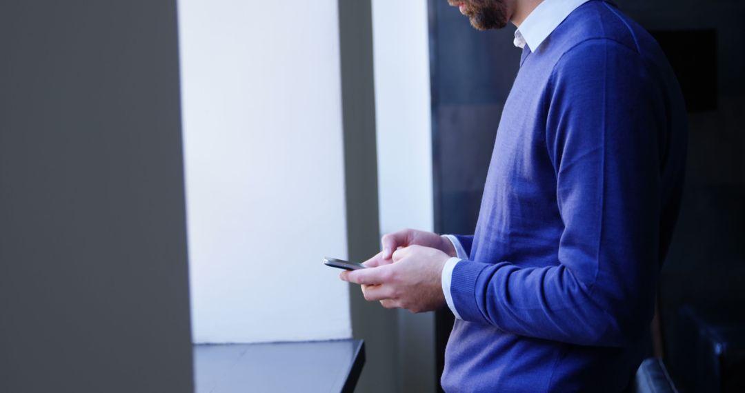 Businessman in Blue Sweater Texting on His Smartphone by Window - Free Images, Stock Photos and Pictures on Pikwizard.com