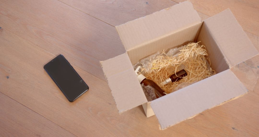 Open Cardboard Box with Packed Bottles and Smartphone on Wood Floor