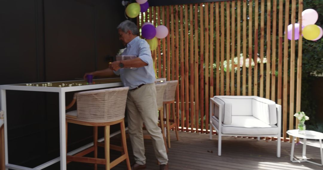 Senior man arranging cups on outdoor bar counter at sunlit wooden patio with balloons