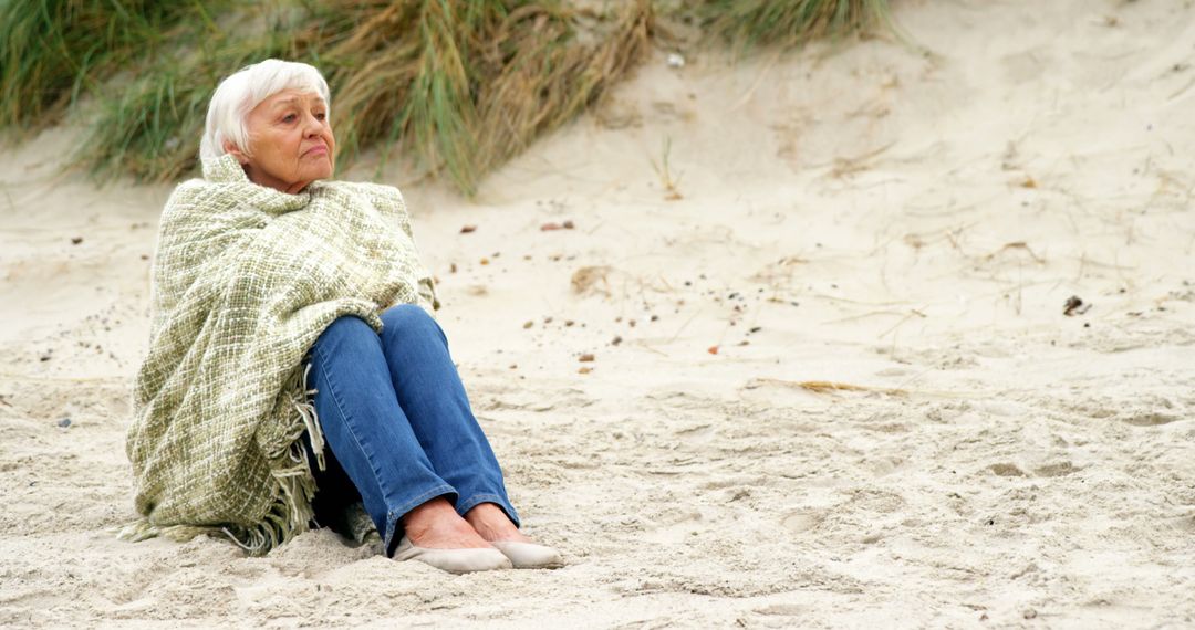 Elderly Woman Sitting on Sandy Beach Wrapped in Blanket - Free Images, Stock Photos and Pictures on Pikwizard.com