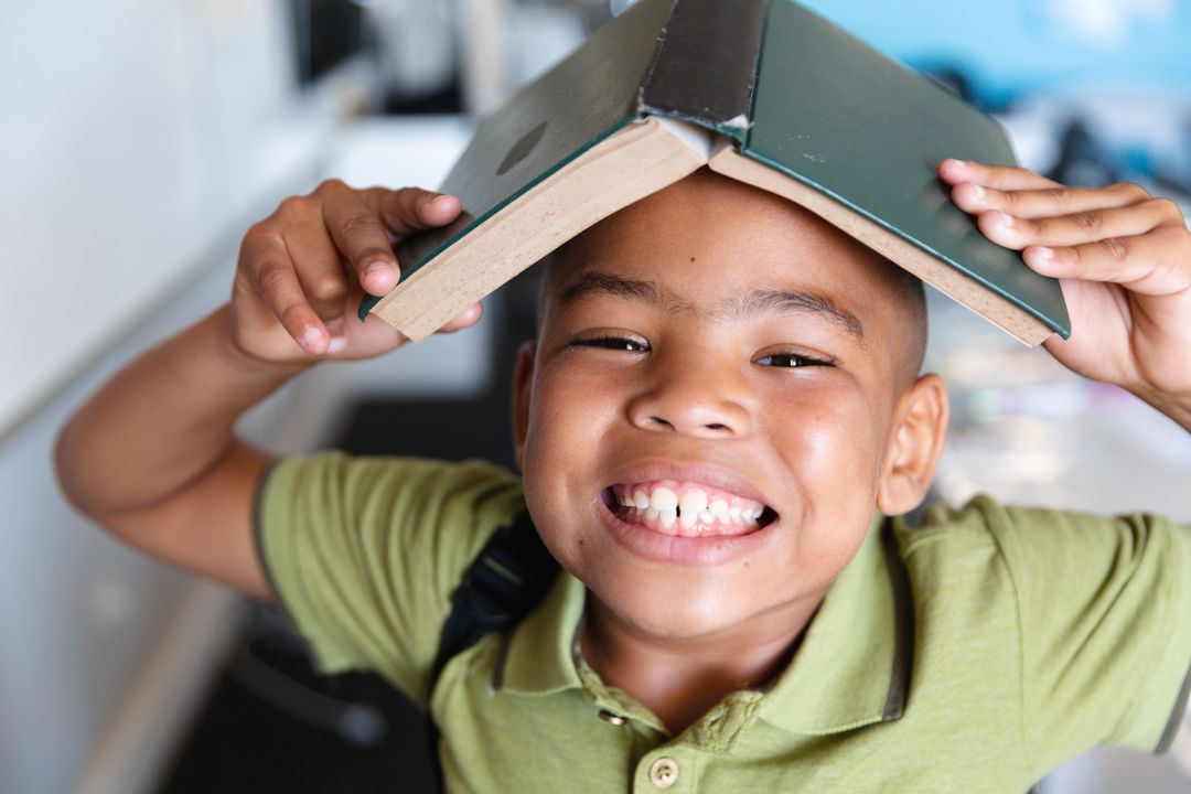 Close-up portrait of smiling african american elementary boy holding book on head in classroom - Free Images, Stock Photos and Pictures on Pikwizard.com