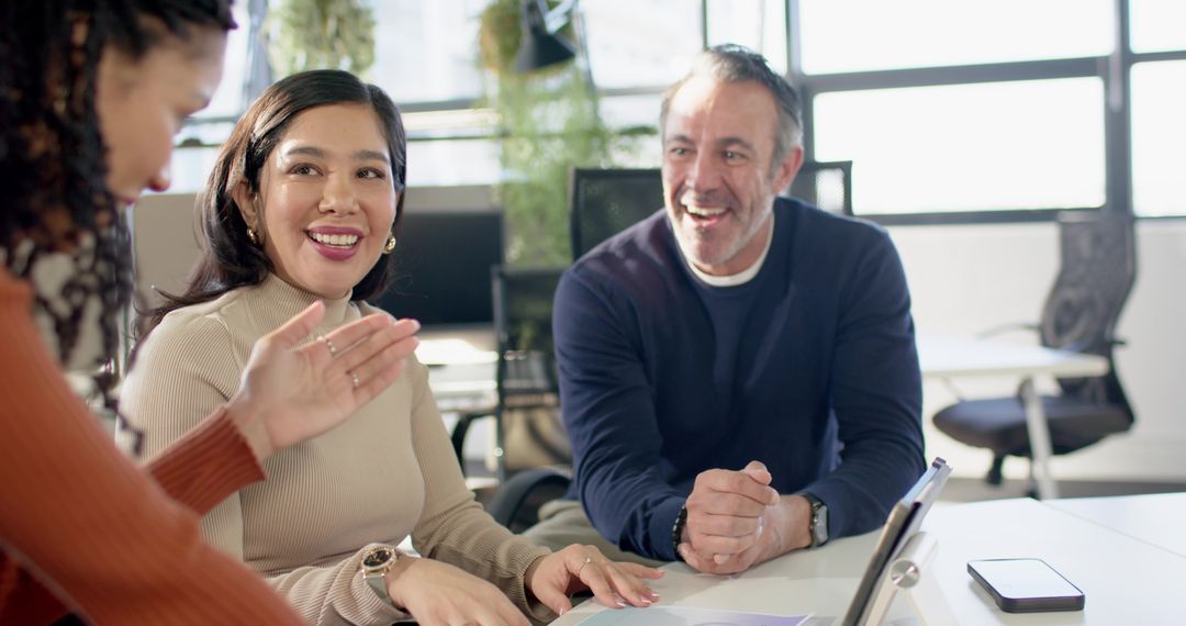 Diverse team collaborating and smiling around laptop in bright modern office with greenery