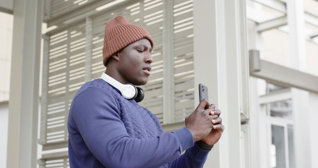 Urban commuter checking smartphone at transit shelter wearing beanie and headphones