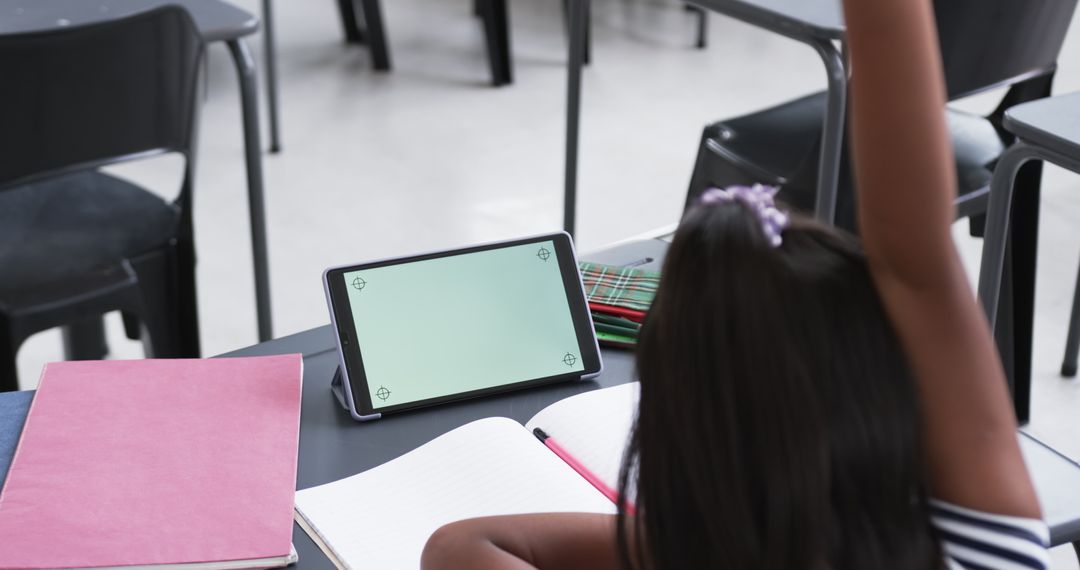 Asian Girl Raising Hand in Classroom Next to Tablet and Books