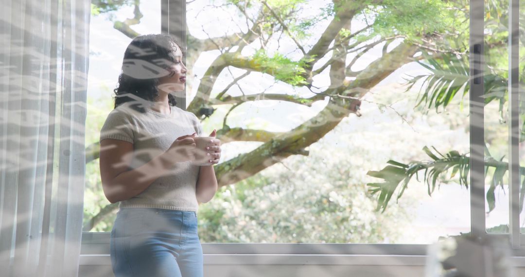Woman holding mug gazing through multi-pane window at leafy tree in soft morning light