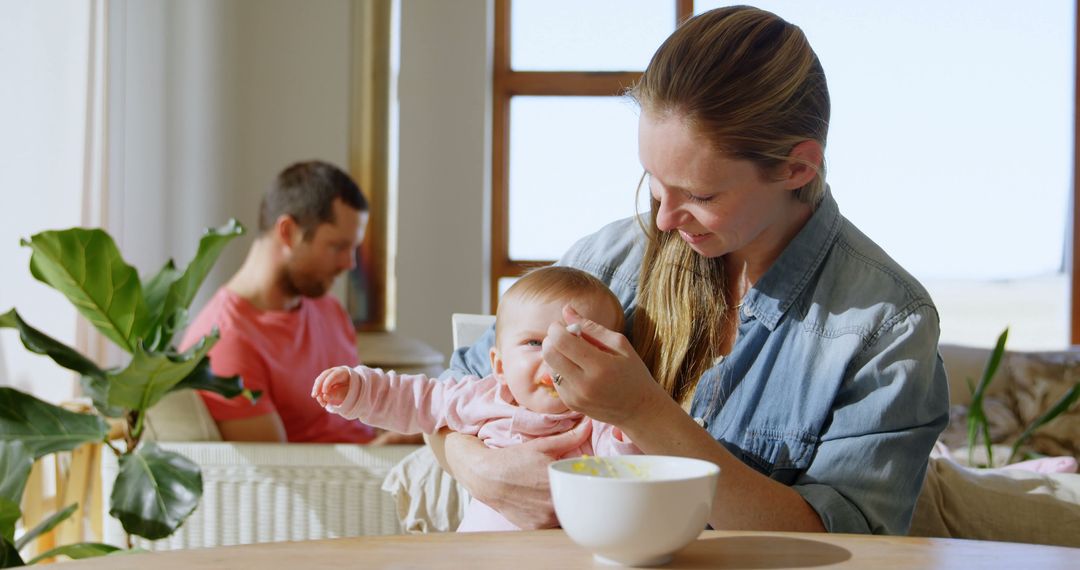 Mother feeding baby in modern living room with father in background - Free Images, Stock Photos and Pictures on Pikwizard.com