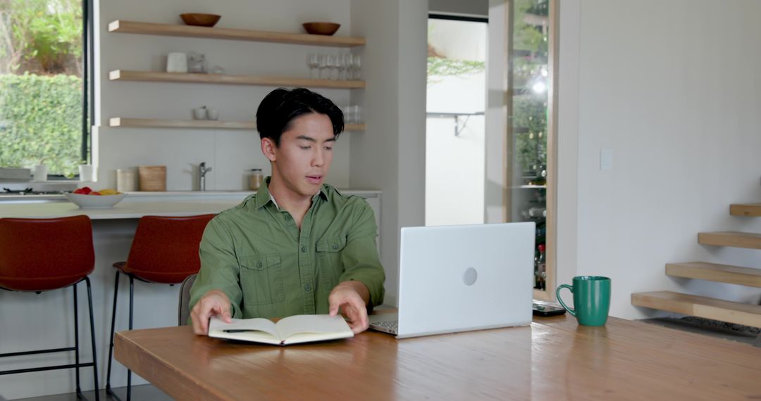 Asian Man Working in Modern Kitchen with Laptop and Book