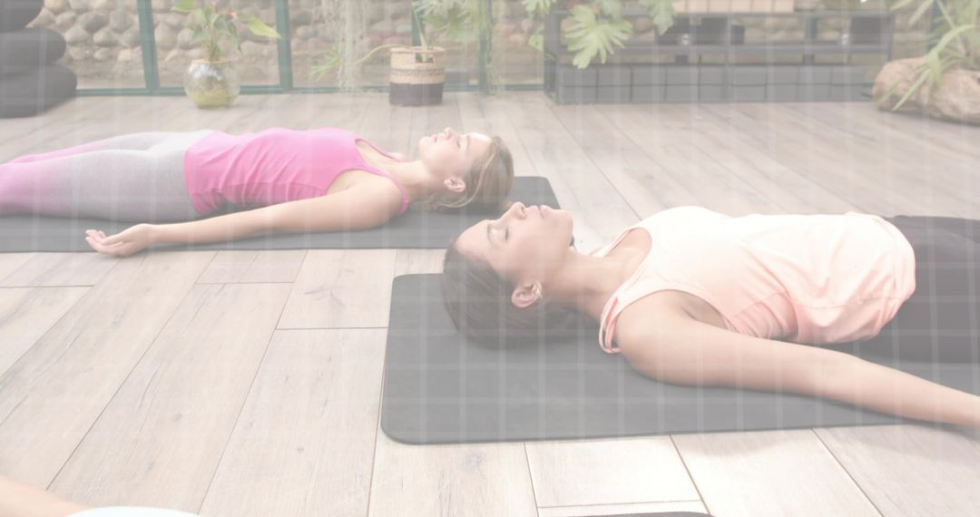 Women practicing supine relaxation yoga on mats in sunlit studio with plants and serenity