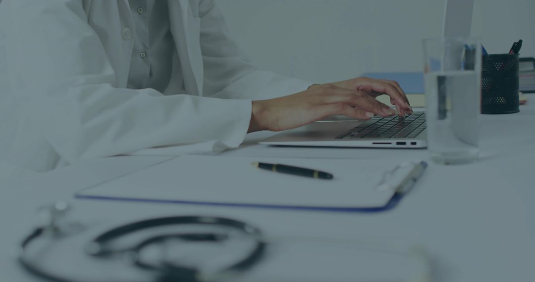 Female Doctor Typing on Laptop at Clinical Desk with Stethoscope and Documents