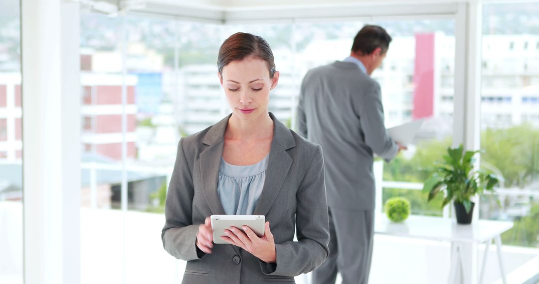 Young Businesswoman Focused on Tablet in Modern Office with Colleague in Background - Free Images, Stock Photos and Pictures on Pikwizard.com