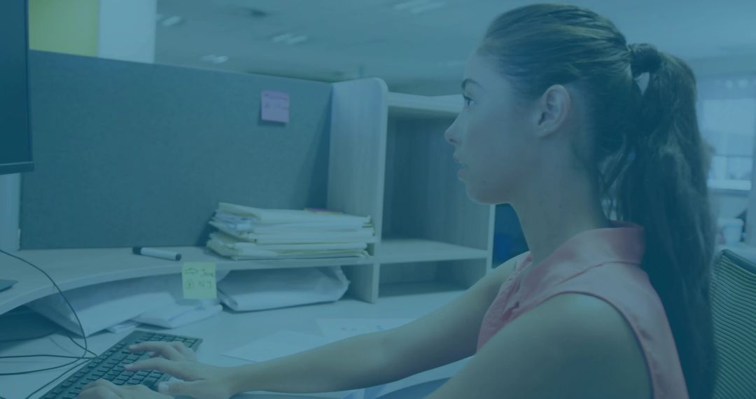 Young woman typing at cubicle desk with monitor, keyboard and stacked files in office