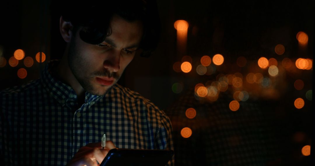 Focused Young Man Using Tablet at Night with City Lights Reflected - Free Images, Stock Photos and Pictures on Pikwizard.com