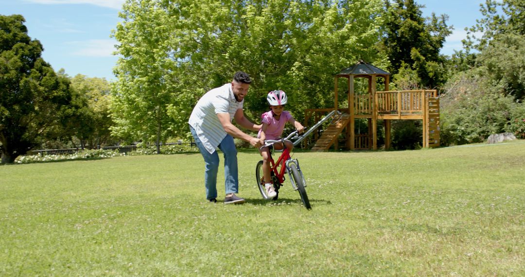 Father Helping Daughter Ride Bicycle in Park Under Blue Sky