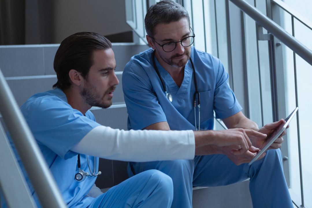 Male surgeons discussing over clipboard while sitting on stairs at hospital - Free Images, Stock Photos and Pictures on Pikwizard.com