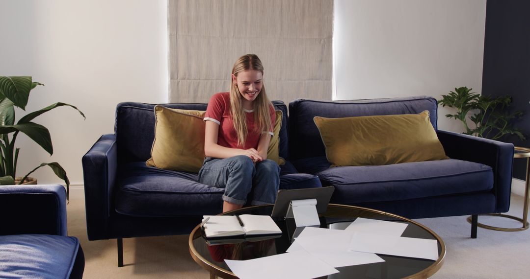 Young woman organizing paperwork while using tablet on navy sofa in cozy modern living room
