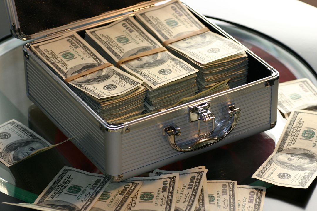Stacks of Cash in Metal Briefcase with Scattered Bills on Glass Table