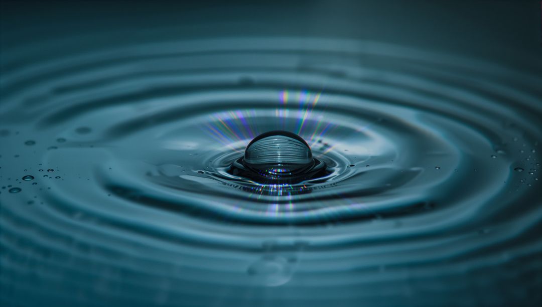 Single Water Droplet Creating Concentric Ripples with Teal Reflections and Diffraction
