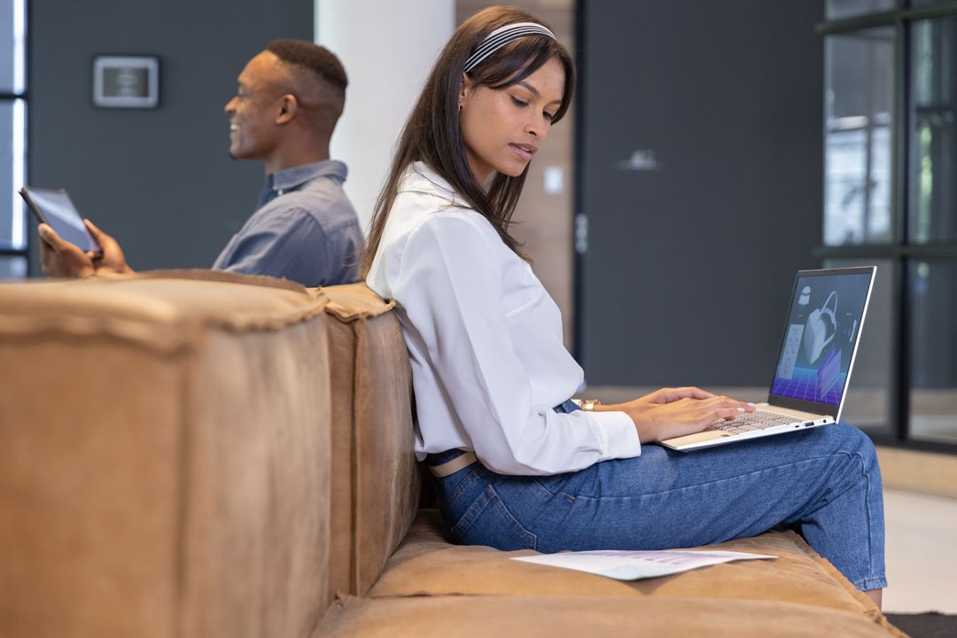 Diverse coworkers working back-to-back on laptop and tablet in modern office lounge