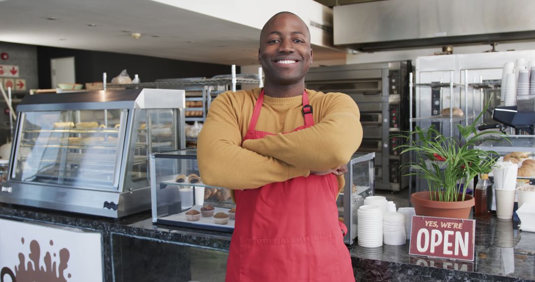 Smiling bakery owner in red apron standing with arms crossed in modern bakery - Free Images, Stock Photos and Pictures on Pikwizard.com