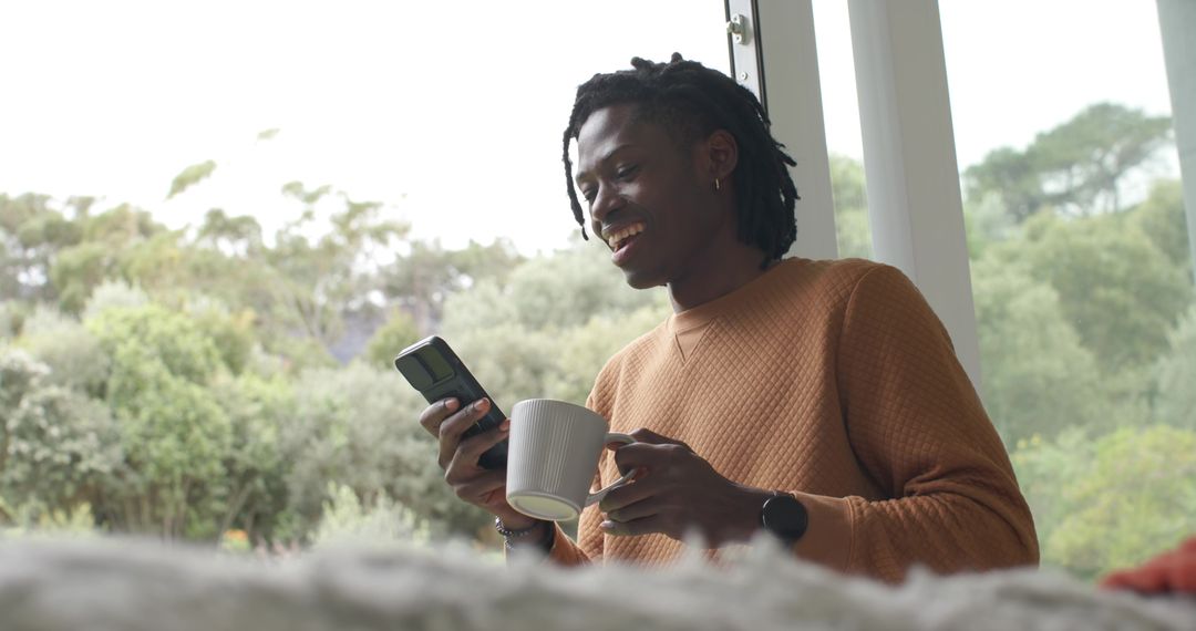 African American Man Enjoying Morning Coffee While Checking Smartphone by Window