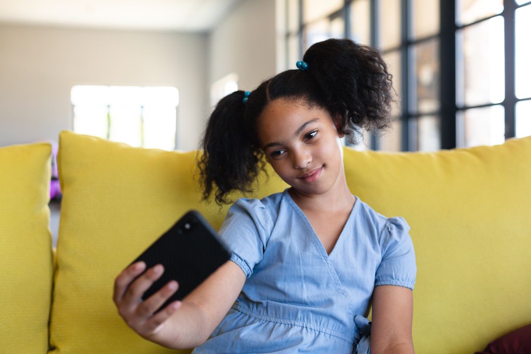 Biracial elementary schoolgirl taking selfie over smart phone while sitting on couch in school - Free Images, Stock Photos and Pictures on Pikwizard.com