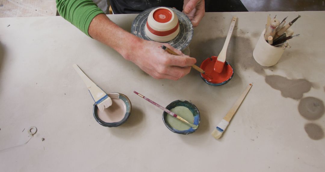 Overhead view of male potter using brush to paint pot on potters wheel at pottery studio - Free Images, Stock Photos and Pictures on Pikwizard.com