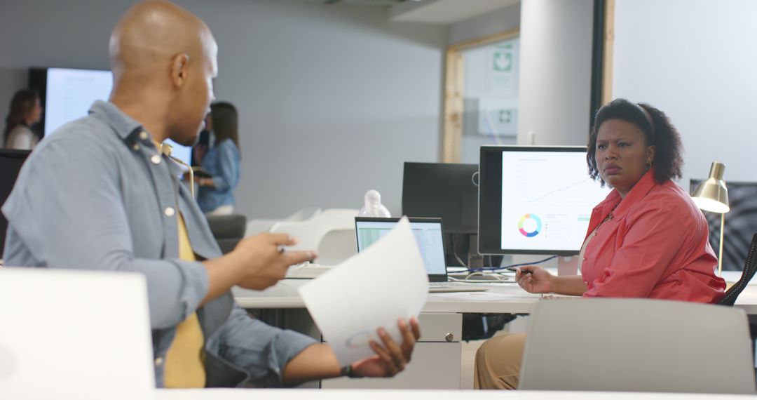 Diverse coworkers debating analytics on screens while holding reports at open office