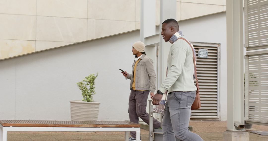 Urban commuters walking with trolley and smartphone along modern building facade, overcast day