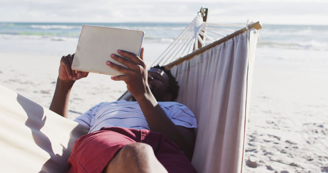 African american man using digital tablet while lying on a hammock at the beach - Free Images, Stock Photos and Pictures on Pikwizard.com