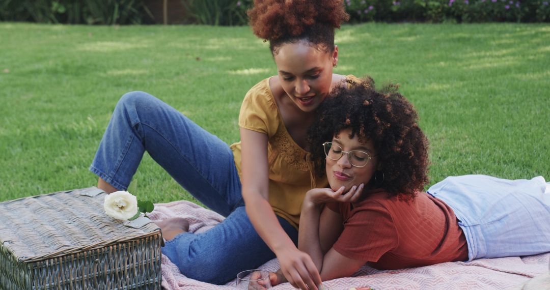 Two women enjoying casual picnic on patterned blanket in park reaching into wicker basket
