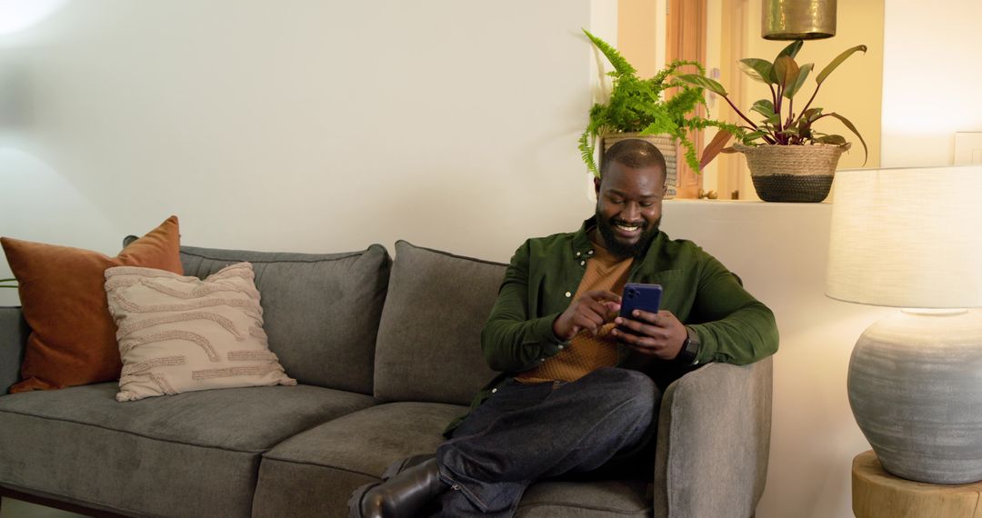 Smiling African American man using smartphone on cozy modern sofa with warm lamp light
