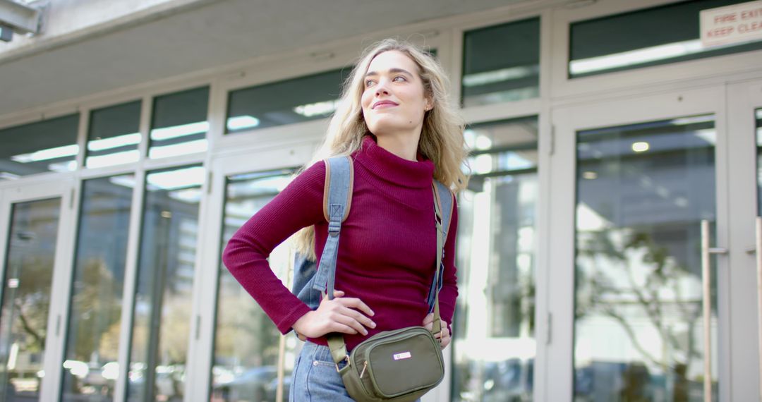 Young woman standing with backpack and olive crossbody bag looking up at glass entrance