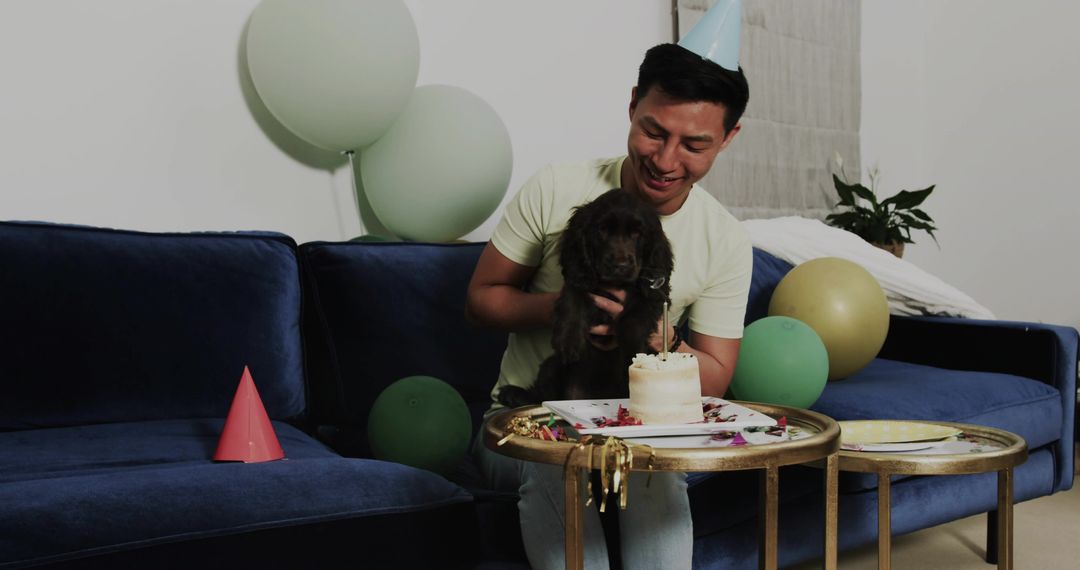 Celebrating Man Holding Puppy and Cake on Blue Sofa with Balloons and Party Hat