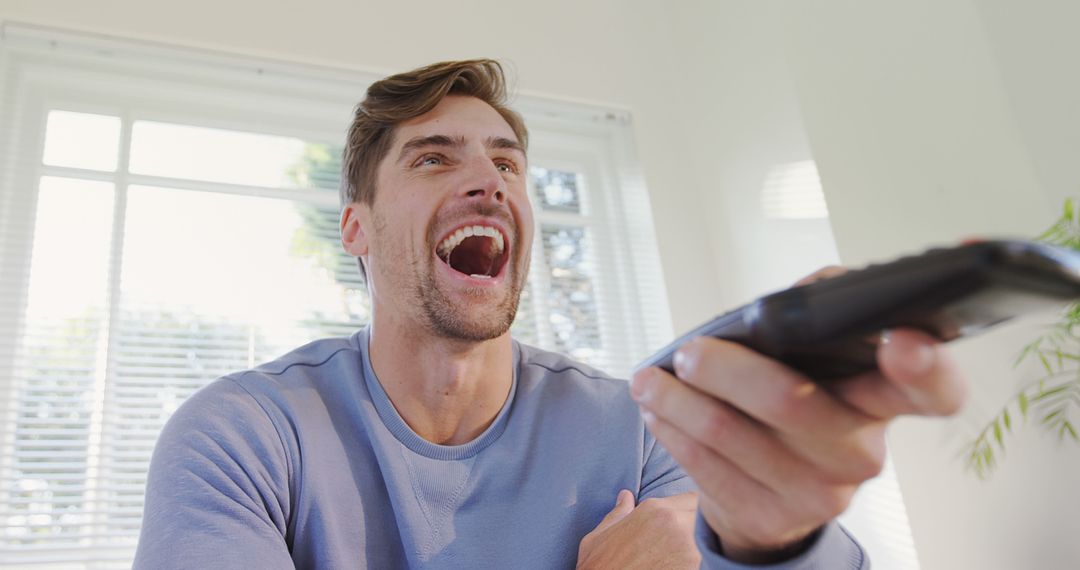 Young Man Laughing and Holding TV Remote in Bright Living Room - Free Images, Stock Photos and Pictures on Pikwizard.com