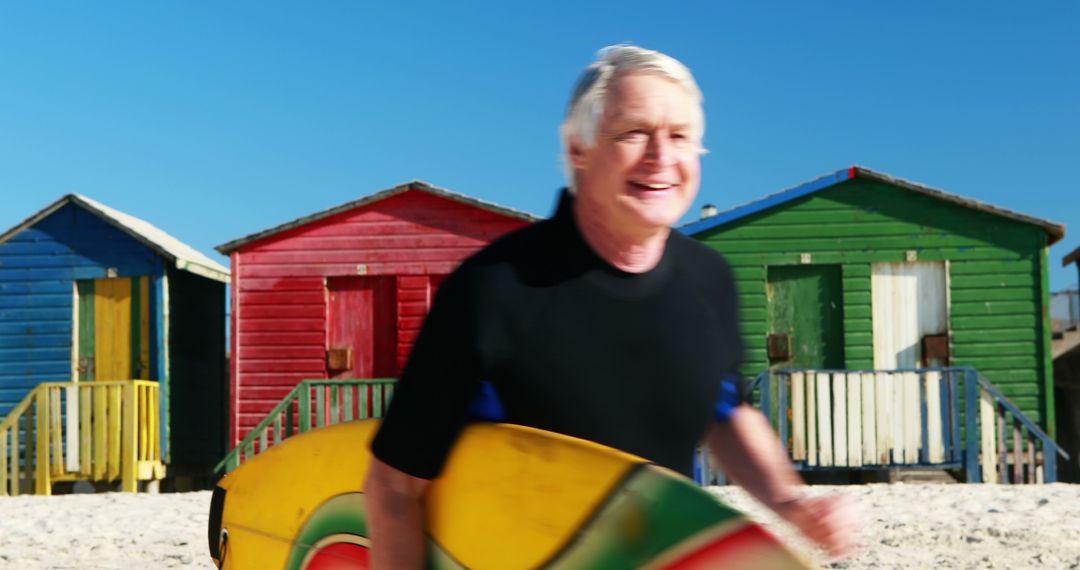 Senior Man Enjoying Beach with Surfboard Near Colorful Beach Huts - Free Images, Stock Photos and Pictures on Pikwizard.com